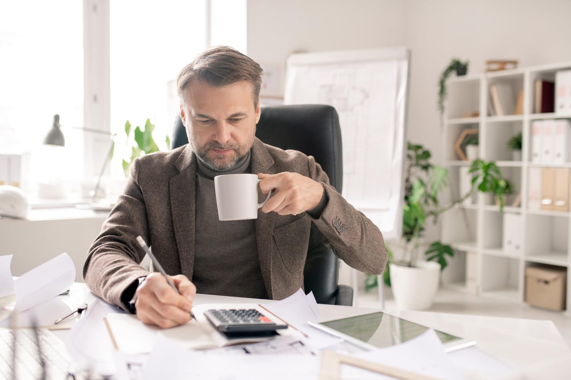 Serious mature accountant with pen and cup of coffee making calculations
